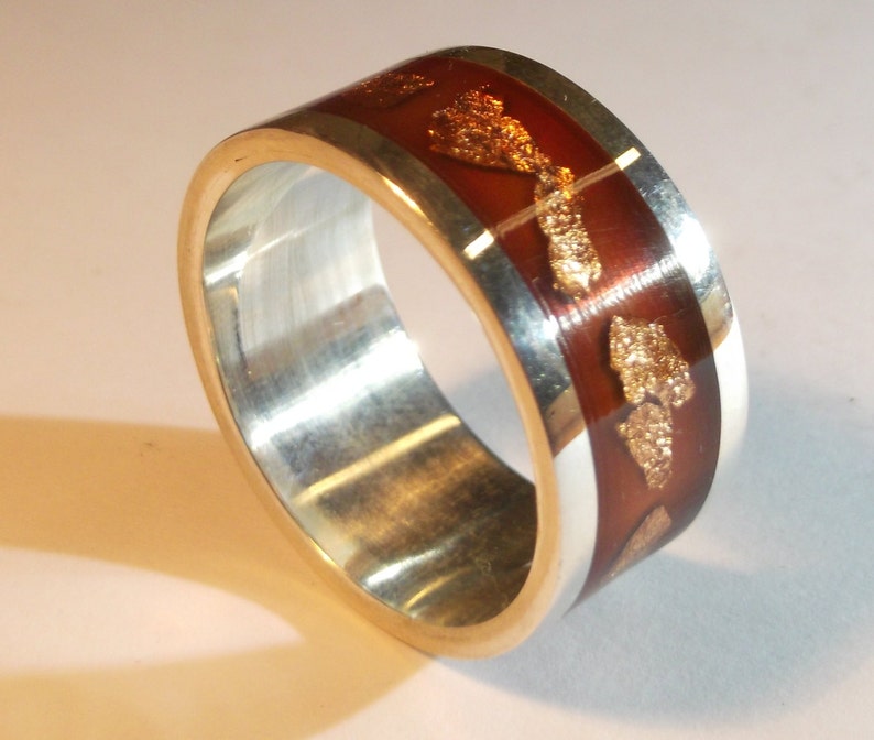 May include: A silver band ring with a brown resin inlay featuring gold flakes. The resin is translucent, allowing the gold flakes to be seen through the brown.