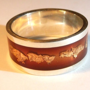 May include: A silver ring with a brown band and gold flakes embedded in the resin. The ring is a wide band and has a polished silver finish.