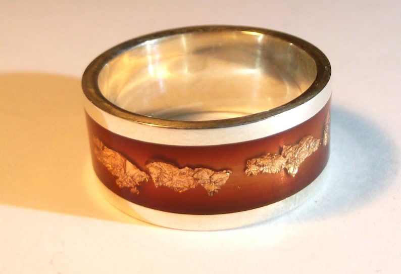 May include: A silver ring with a brown band and gold flakes embedded in the resin. The ring is a wide band and has a polished silver finish.