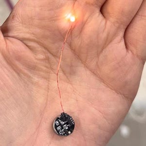 Op de afbeelding: Een kleine, ronde, zwarte printplaat met een zilveren rand, verbonden met een dunne rode draad en een helder oranje licht. Het licht is ingeschakeld, wat suggereert dat het apparaat is ingeschakeld. De componenten worden in een hand gehouden.