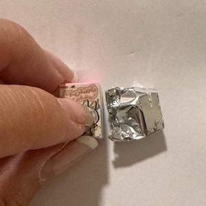 May include: A small, pink and white box with Japanese text and illustrations is held next to a silver foil-wrapped cube. The box has a decorative design. The foil-wrapped cube is a square shape.