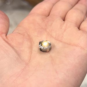 Könnte beinhalten: Eine kleine, runde LED-Leuchte mit hellem, gelb-weißem Leuchten. Sie hat ein silberfarbenes Metallgehäuse und wird in einer Hand gehalten. Diese Miniaturleuchte ist ein Bauteil, wahrscheinlich für Elektronik oder Bastelarbeiten.