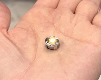 May include: A small, round LED light with a bright, yellow-white glow. The light has a silver-colored metal casing and is held in the palm of a hand. The light is a miniature lighting component.