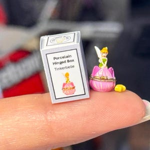 Könnte beinhalten: Eine Miniatur-Porzellan-Scharnierbox mit der Aufschrift "Tinkerbelle" und einer passenden Illustration. Neben der Box befindet sich eine kleine rosa-goldene Schmuckschatulle mit einer Feenfigur. Ein gelber sternförmiger Anhänger ist ebenfalls vorhanden.