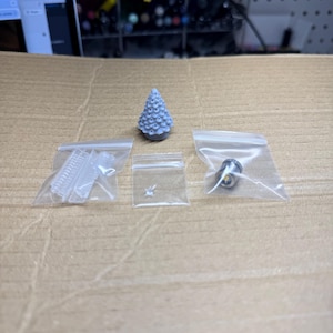 May include: A small gray 3D printed Christmas tree with a white LED light, a small white plastic piece, and a silver metal piece in separate plastic bags.