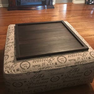 May include: A square ottoman with a dark brown wooden tray on top. The ottoman is covered in a cream fabric with a repeating pattern of black text and floral designs. The text includes words like "The", "of Menthe", "Provence", "Darjeeling", and "Ceylan".