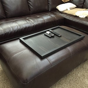 May include: A black wooden tray with a dark wood grain finish sits on top of a brown leather ottoman. The tray is holding several remote controls.