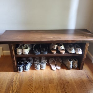 May include: A dark brown wooden shoe rack with two shelves. The top shelf has a variety of shoes, including sandals, sneakers, and flats. The bottom shelf has a variety of sneakers and flats.