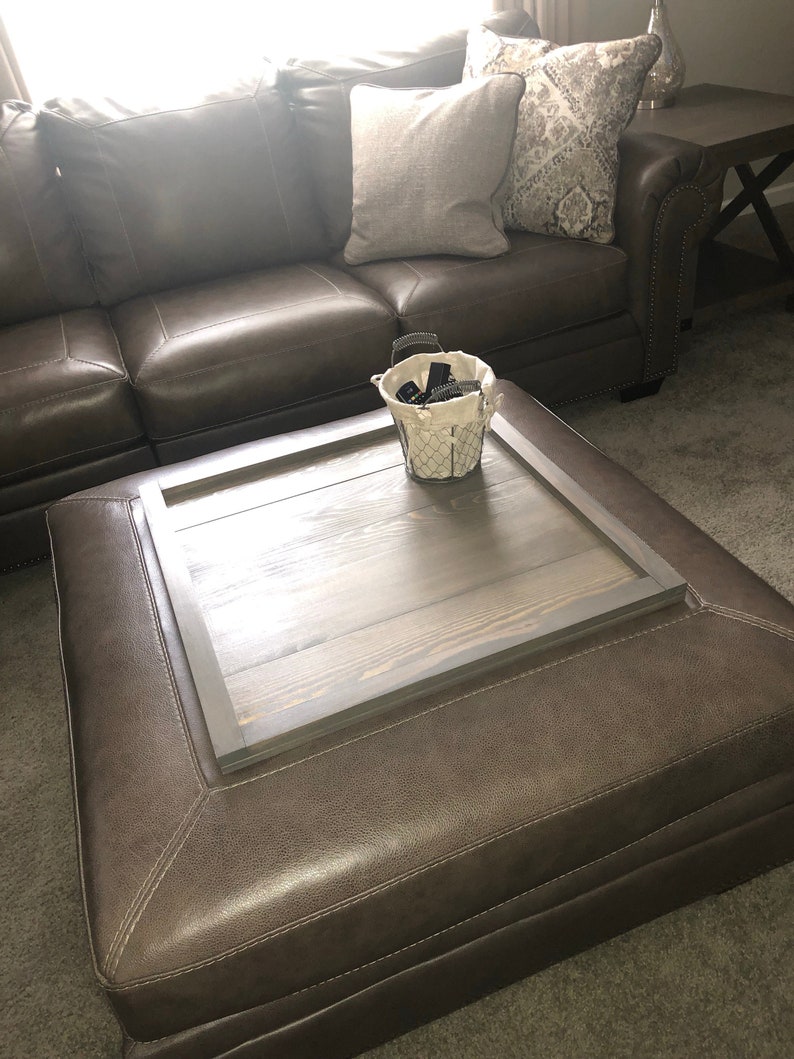 May include: A brown leather ottoman with a wooden tray top. The tray has a dark gray frame and is perfect for holding items like remotes or books.