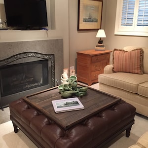 May include: A brown leather ottoman with a wooden top and a book titled "The Joy of the River" on top. The ottoman is in a living room with a couch, fireplace, and a lamp on a wooden table.