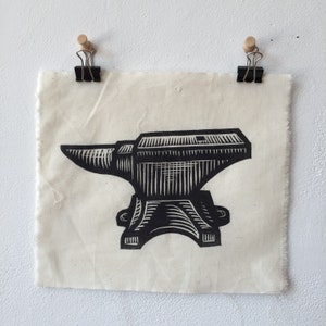 Op de afbeelding: Een zwart-wit linoleumsnede van een aambeeld. De print is bevestigd aan een stuk ecru stof met twee zwarte klemmen en houten punaises. Het aambeeld is een traditioneel smidsgereedschap.