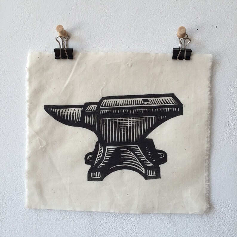May include: A black and white linocut print of an anvil. The print is attached to a piece of off-white fabric with two black binder clips and wooden drawing pins. The anvil is a traditional blacksmithing tool.