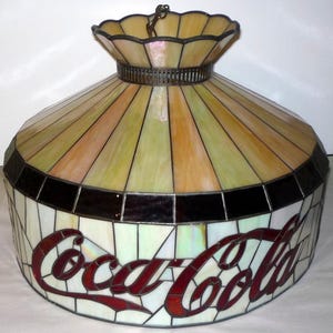 May include: A stained glass Coca-Cola lamp shade. The shade features a multi-colored design with the Coca-Cola logo in red. The top of the shade has a scalloped edge and a metal ring for hanging. The shade is a mix of yellow, orange, and green.