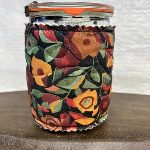 May include: A clear glass jar with a metal clamp lid and an orange silicone seal, encased in a quilted fabric sleeve featuring a black background with a repeating pattern of stylized flowers in shades of orange, red, and green.