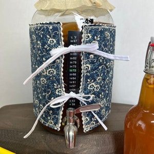 May include: A glass jar with a blue and white floral fabric cover, a white ribbon tied around the top, and a stainless steel spigot. The jar has a thermometer attached to it, with a reading of 22.72 degrees Celsius. A clear glass bottle with a brown liquid is in the background.