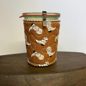 May include: A glass jar with a lid and metal clasp, wrapped in a brown fabric sleeve. The sleeve is patterned with white cowboy boots. The jar sits on a dark wooden surface.