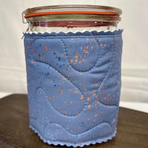 May include: A glass jar with a metal clamp lid, encased in a quilted fabric cozy. The cozy is a blue color with orange and blue speckles. The jar is sitting on a wooden surface.