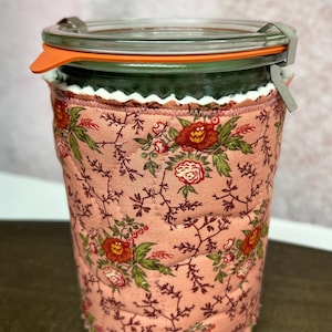 May include: A glass jar with an orange lid and a floral fabric cozy. The cozy is a quilted peach fabric with red and green floral patterns. The jar has a metal clasp and a white scalloped trim.