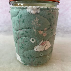 May include: A glass jar with a red-orange rubber seal and a metal clasp, encased in a quilted, light green fabric cozy. The cozy features a white scalloped edge and a pattern of white rabbits and floral designs.