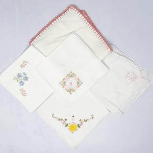 May include: A collection of white handkerchiefs with embroidered floral designs. One handkerchief has a pink crocheted edge. The embroidery features blue, pink, yellow, and green colors. The handkerchiefs are arranged on a white surface.