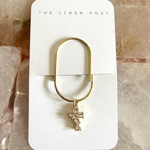 May include: Gold-toned metal paper clip with a dangling cross pendant. The cross is adorned with clear, sparkling gemstones. The paper clip is attached to a white card with the text "THE LINEN POST" printed at the top.