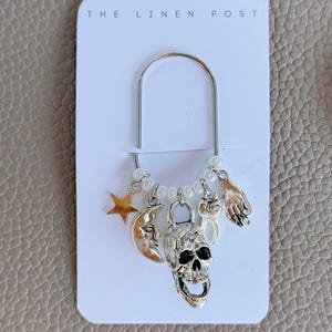 May include: A silver-toned metal paperclip charm holder on a white card labeled THE LINEN POST. The clip features dangling charms including a gold star, a silver crescent moon, a skull, a white tooth, and a silver hand, accented with small white beads.