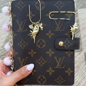 May include: A brown planner with a repeating monogram pattern, featuring a gold-tone chain with light pink pom-poms, a gold-tone paperclip bookmark with a seashell and bow charm, and a second gold-tone paperclip bookmark with a seashell charm.