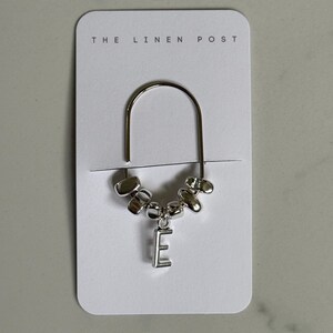 May include: A silver bookmark with a curved top and a dangling charm. The charm includes the letter "E" and several small, rounded beads. The bookmark is attached to a white card with "THE LINEN POST" printed at the top.