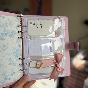 A hand holding a pink notebook with a clear pocket, a pink ribbon, and a white card with text on it.