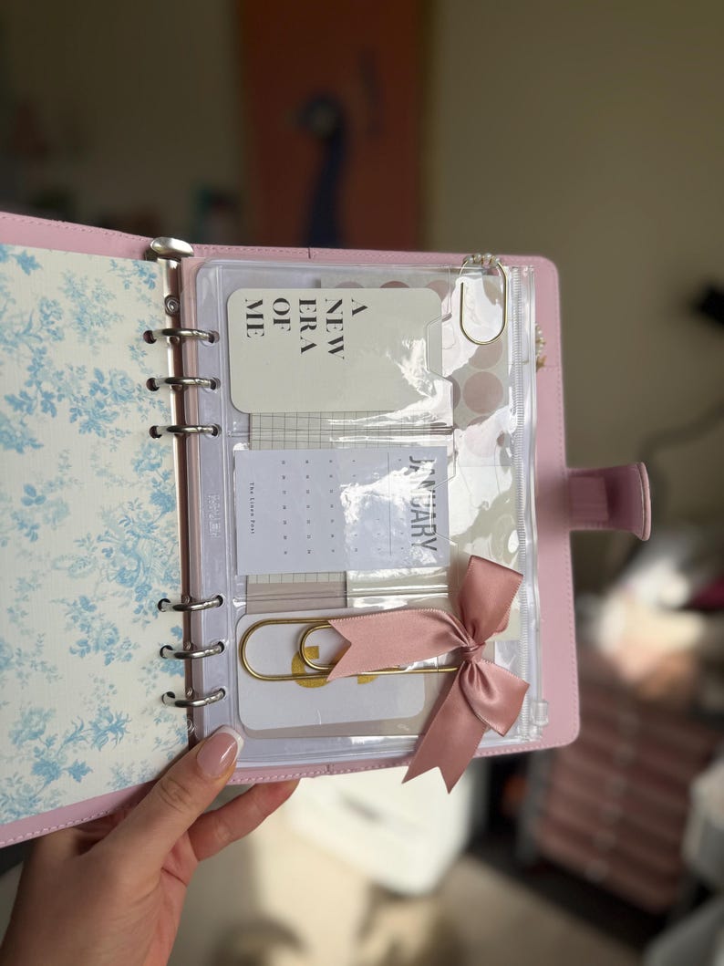 A hand holding a pink notebook with a clear pocket, a pink ribbon, and a white card with text on it.