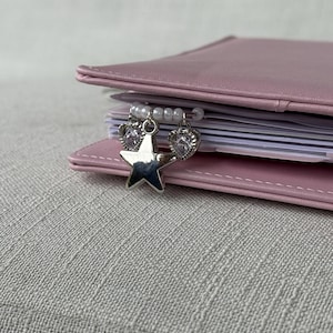 Puede incluir: Una agenda de cuero rosa con un dije de estrella y corazón plateado colgando de una cadena de cuentas. La agenda está abierta, revelando páginas blancas. El dije está sujeto a la encuadernación de la agenda.