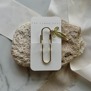 May include: A gold-colored paper clip with a decorative bow detail, presented on a white card with the text "THE LINEN POST". The clip is displayed on a textured, light-colored stone, with white and beige fabric accents.