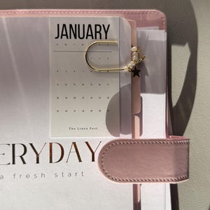 May include: A gold-toned metal paperclip bookmark adorned with small white pearls and a tiny gold star charm, clipped onto a January calendar page inside a light pink planner with the text EVERYDAY a fresh start visible on the cover.