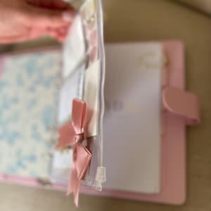 A hand holding a pink binder with a clear zipper, and a white paper inside the binder.