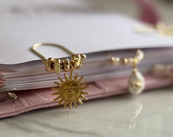 Könnte beinhalten: Ein goldfarbener Sonnenanhänger mit einem lächelnden Gesicht hängt an einer goldfarbenen Sicherheitsnadel. Der Anhänger ist an einem rosa Leder-Notizbuch befestigt. Der Hintergrund ist verschwommen, scheint aber ein Schreibtisch oder Tisch zu sein.
