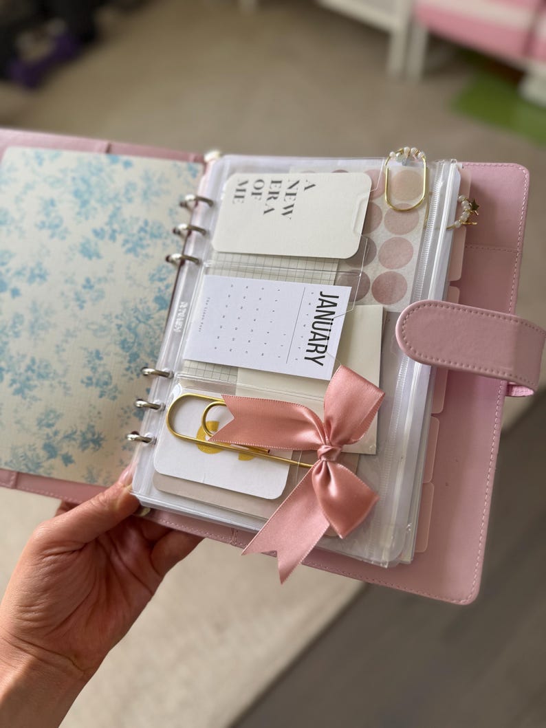 A person's hand holding a pink and white spiral-bound notebook with various items such as a calendar, a ribbon, and other small items.