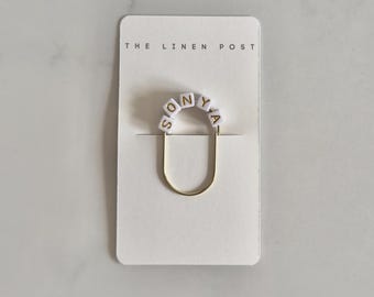 May include: A gold-toned U-shaped metal clip featuring five white square beads with gold lettering that spell out SONYA. The clip is attached to a rectangular white display card with the text THE LINEN POST printed at the top in black.