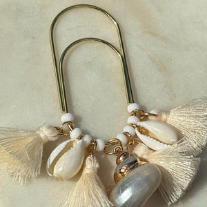 May include: A gold-colored paperclip adorned with seashells, white beads, and cream-colored tassels. The seashells are in shades of white and beige, with a pearlescent sheen on one. The tassels add a delicate touch to this unique accessory.