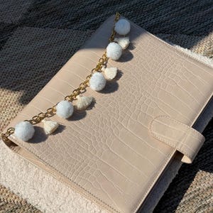 May include: A beige rectangular planner with a crocodile-embossed texture and a side tab closure. A gold-toned chain is attached to the top, featuring alternating white fuzzy pom-poms and small cream-colored fabric tassels as decorative accents.