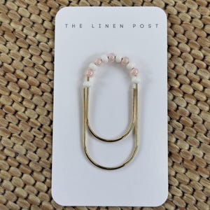 May include: A gold-coloured paper clip with a beaded accent. The beads are white and pink, arranged in an arch shape. The paper clip is on a white card with the text "THE LINEN POST". The background is a woven texture.