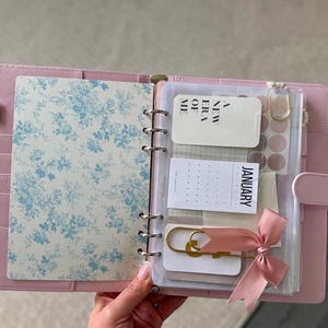 A person's hand holding a pink-colored spiral notebook with a floral pattern, which appears to be a personal organizer or planner.
