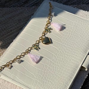 May include: A white rectangular notebook with a textured crocodile skin pattern featuring a gold-toned chain charm attached to the cover. The charm includes a smooth gold heart, two small light pink tassels, and several small floral accents.