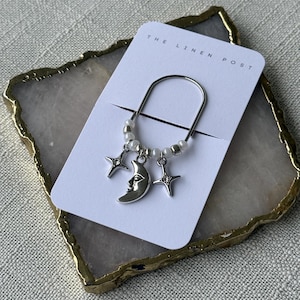 May include: A silver-toned bookmark with a crescent moon charm and two star charms, accented with small white beads. The bookmark is attached to a white card with the text "THE LINEN POST". The card rests on a decorative, gold-edged stone.