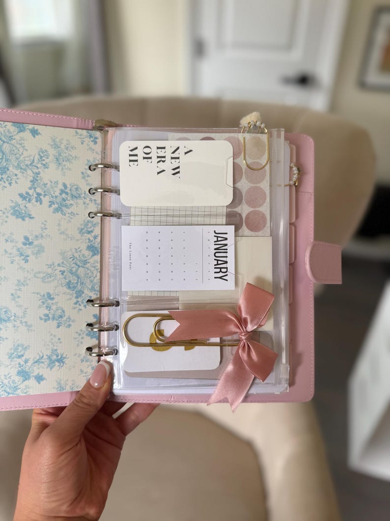 A person's hand holding a pink and white spiral-bound notebook with various items such as a calendar, a pen, and a pink ribbon.