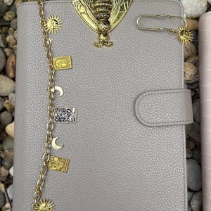 May include: A light gray textured notebook featuring a large gold bee emblem at the top. A gold chain drapes down the left side, adorned with charms including suns, moons, and rectangular tarot-style cards. A gold sun charm is attached to a paperclip.