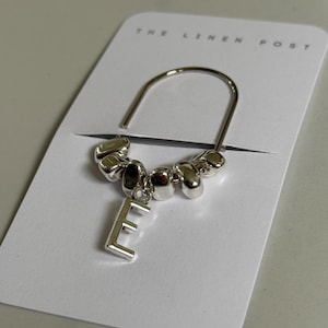 May include: A silver-toned bookmark with a curved top and a dangling letter "E". The bookmark features a series of small, rounded beads. The bookmark is displayed on a white card with the text "THE LINEN POST".