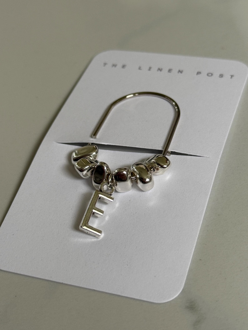 May include: A silver-toned bookmark with a curved top and a dangling letter "E". The bookmark features a series of small, rounded beads. The bookmark is displayed on a white card with the text "THE LINEN POST".