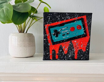 May include: A black canvas painting with a red and blue video game controller dripping red paint. The background is black with white splatter paint.