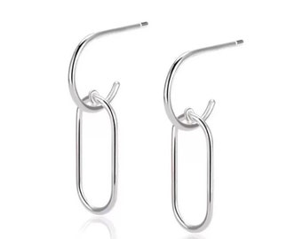 May include: A pair of silver-colored hoop earrings with a dangling oval-shaped charm. The earrings are made of a shiny, reflective metal and are displayed against a white background. The design is minimalist and modern, suitable for everyday wear.