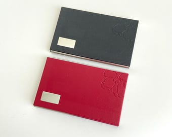 May include: Two business card holders, one black and one red, with a silver accent and a floral design.